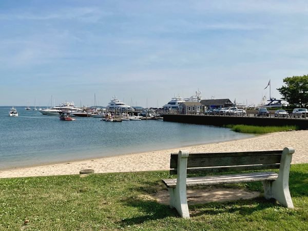 A bench on the lawn by the beach in Sag Harbor, boats at the pier behind